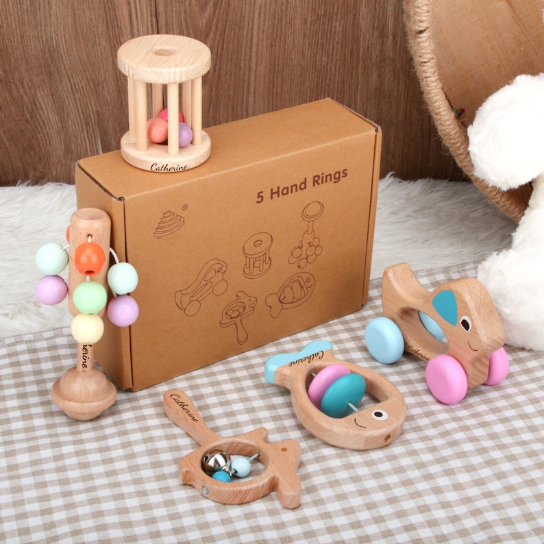 May include: A collection of wooden baby toys, including a rattle, a rolling cylinder, and a car-shaped toy. The toys are natural wood with colorful accents and are displayed with a box that reads "5 Hand Rings".