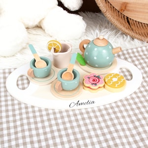 Può includere: Un set da tè giocattolo in legno su un vassoio ovale bianco. Il set include una teiera turchese a pois bianchi, due tazze, piattini, cucchiai e biscotti. Il vassoio è personalizzato con il nome "Amelia".