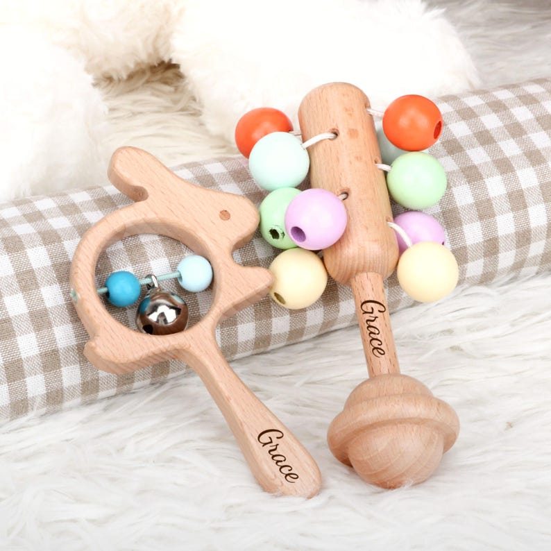 May include: Two wooden baby rattles. One is shaped like a bunny with a bell and colorful beads. The other is a mallet shape with colorful beads. Both rattles have the name "Grace" engraved on them.