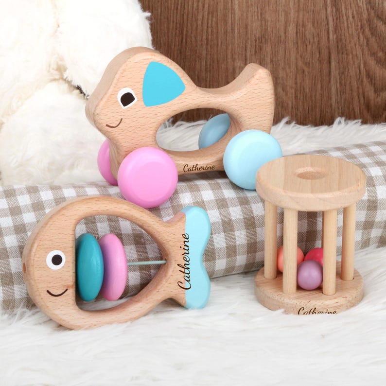 May include: Wooden baby toys, including two fish-shaped rattles with blue and pink accents, and a small wooden cage with colorful balls inside. The name "Catherine" is printed on the toys.