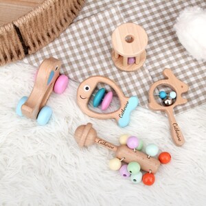 May include: A collection of wooden baby toys, including a pull-along elephant, a fish-shaped rattle, a bunny-shaped rattle, a drum-shaped rattle, and a beaded rattle. The toys are made of light-colored wood with pastel-colored accents and the name "Catherine" is engraved on some of them.