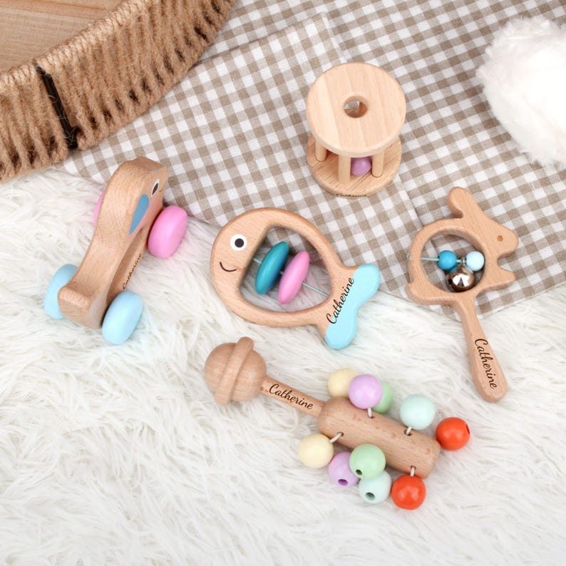 May include: A collection of wooden baby toys, including a pull-along elephant, a fish-shaped rattle, a bunny-shaped rattle, a drum-shaped rattle, and a beaded rattle. The toys are made of light-colored wood with pastel-colored accents and the name "Catherine" is engraved on some of them.