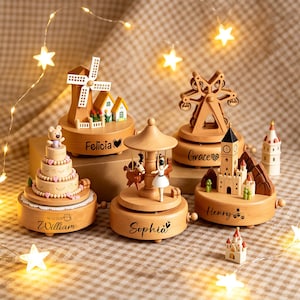 May include: Five wooden music boxes featuring various designs including a windmill, a tiered cake with a bear, a carousel with a dancer, a Ferris wheel, and a clock tower. Each base is engraved with names like Felicia, William, Sophia, Grace, and Henry.