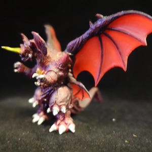 May include: A miniature fantasy figurine of a dragon with red wings, purple accents, and a yellow horn. The dragon has a detailed, textured appearance and is posed against a dark background.