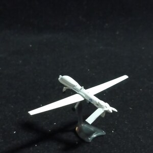 May include: A small, white model airplane with long wings and a detailed body. The model is mounted on a black stand, and the wings are outstretched. The model airplane is a miniature replica of a drone.