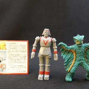 May include: Three vintage Japanese robot and monster figurines. The robot is gray with red accents, the monster is teal with gold details, and a card with Japanese text is on the left.