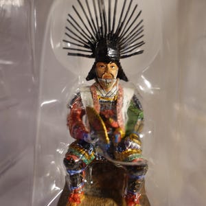 Puede incluir: Una figurita colorida de un guerrero samurái japonés que lleva un casco negro con una corona puntiaguda y una armadura adornada. La figurita está sentada sobre una base de madera.