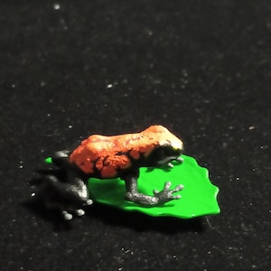 Puede incluir: Una figura de rana en miniatura con un cuerpo naranja y negro vibrante, posada sobre una hoja verde brillante. Los detalles de la rana y el acabado brillante de la hoja crean un contraste llamativo contra el fondo oscuro.