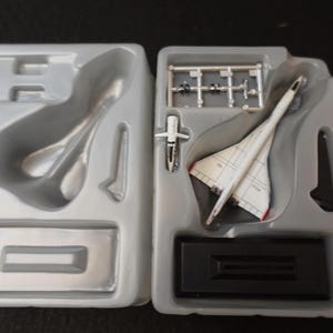 Pode incluir: Kit de modelo de avião Concorde branco e vermelho. O kit inclui o modelo do jato, um pequeno avião e peças adicionais, tudo embalado numa bandeja de plástico cinzento. O modelo é exibido numa base preta.