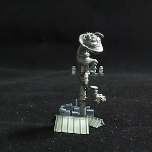 May include: A detailed miniature sculpture of a figure perched atop a structure resembling a radio tower. The figure is gray, with a large hat. The tower has a cityscape base and various mechanical components. The overall color palette is muted, with a focus on gray and white.