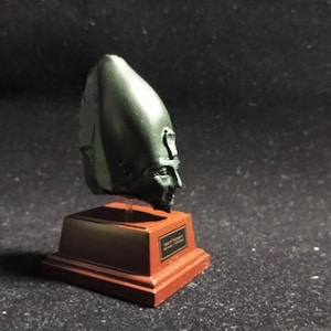 Könnte beinhalten: Eine dunkelgrüne Büste eines Pharaos, montiert auf einem rechteckigen, rotbraunen Sockel. Die Büste weist detaillierte Gesichtszüge und eine hohe Kopfbedeckung auf. Der Sockel hat eine goldfarbene Plakette mit Text. Die Skulptur wird vor dunklem Hintergrund gezeigt.