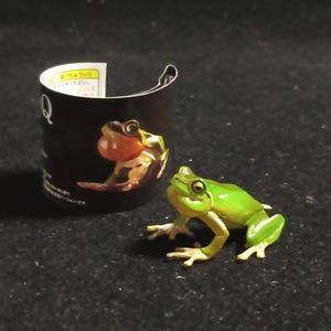 May include: A small, green frog figurine with a light green body and yellow feet. The frog is positioned next to a black cylinder with a frog illustration. The frog is on a dark surface.