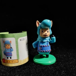 May include: A small, blue figurine of a character with animal-like features, wearing a blue outfit with red accents. The figurine stands on a green base. A small, illustrated paper container is next to the figurine, with Japanese text and a matching character image.