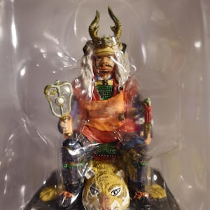 Puede incluir: Una figurita de un guerrero samurái con un casco con cuernos y una armadura adornada, sentado sobre un tigre. El guerrero sostiene un cetro ceremonial en su mano derecha. La figurita está pintada en colores vibrantes, incluyendo rojo, dorado y verde.
