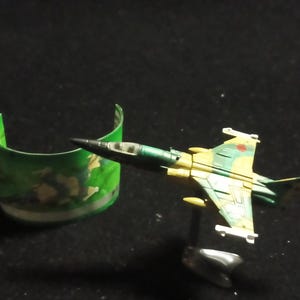 Könnte beinhalten: Ein Miniatur-Spielzeug-Kampfjet in Grün-, Gelb- und Brauntönen mit einer schwarzen Nase. Der Jet steht auf einem silbernen Ständer. Daneben befindet sich ein grün-weißes, gebogenes Objekt mit Tarnmuster.