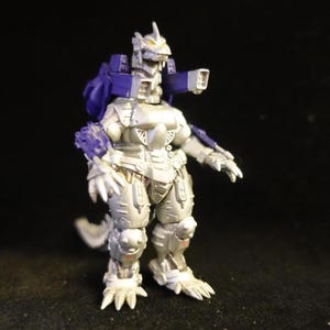 May include: A silver and purple robotic action figure, possibly a collectible toy. The figure has a detailed design with articulated joints, a spiked tail, and a head with a distinctive shape. The figure is standing on a dark surface.