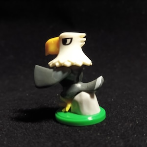 Peut inclure: Petite figurine d'aigle avec une tête blanche, un bec jaune et des ailes grises. L'aigle est perché sur un rocher gris sur une base circulaire verte. Le fond est sombre.