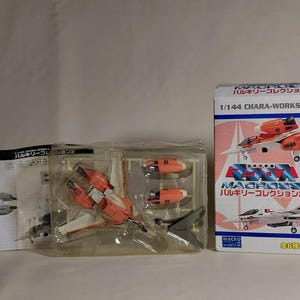 Puede incluir: Un kit de modelo de un caza Valkyrie Macross, con una combinación de colores naranja y blanco. El kit incluye varias piezas y está empaquetado con una caja que muestra el avión y texto japonés.