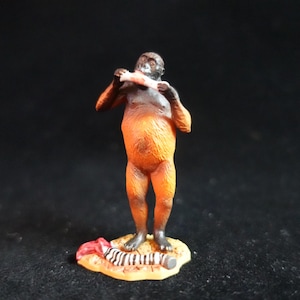 May include: A small orange and black figurine of a primate holding a bone in its hands. The figurine is standing on a small base with a red and white striped pattern.