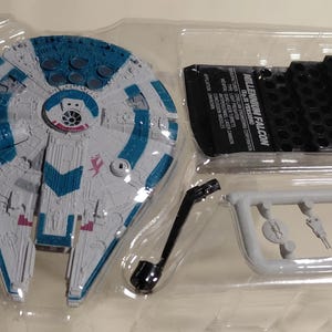 May include: A Millennium Falcon model kit in a clear plastic package. The model is primarily gray with blue and red accents. The kit includes various parts, including a black stand and a sprue of smaller components. The text "MILLENNIUM FALCON" is visible.
