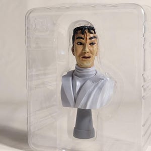 May include: A bust of a man with a scar on his forehead, wearing a white turtleneck and a light gray jacket. The bust is in a clear plastic package. The figure has a neutral expression.