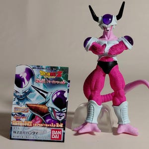 Puede incluir: Figura de acción rosa y blanca de un personaje de Dragon Ball Z con una caja con texto japonés. La figura tiene una constitución muscular, una larga cola y cuernos negros. La caja muestra imágenes del personaje.