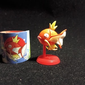 Könnte beinhalten: Eine orangefarbene und weiße Magikarp-Figur auf einem roten Ständer, neben einem blau-roten zylindrischen Behälter mit einer Magikarp-Illustration. Die Figur hat eine gelbe Flosse und einen überraschten Gesichtsausdruck. Der Behälter hat eine Magikarp-Illustration und japanischen Text.