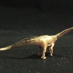 May include: A tan dinosaur figurine with a long neck and tail. The figurine has a textured surface and is posed on all fours. The dinosaur has a row of small spikes along its back.