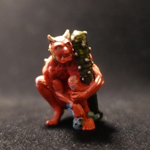 May include: A red and gold figurine of a demon holding a green and gold staff. The demon has horns and a tail.