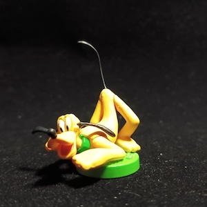 May include: A yellow Pluto figurine with a black nose and ears, green collar, and a black tail. The figurine is lying down on a green circular base. The background is black.