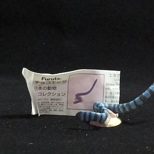 Puede incluir: Una pequeña y detallada figura de una serpiente azul y negra, enrollada sobre una pequeña base beige. Un papel blanco con texto japonés está junto a la serpiente. La figura es parte de una colección.