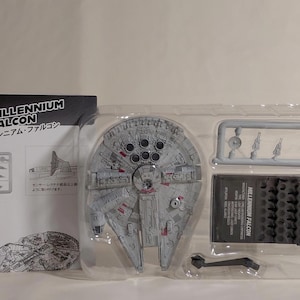 Może przedstawiać: Zestaw modelu Millennium Falcon w przezroczystym plastikowym opakowaniu. Model jest szary z czerwonymi i czarnymi akcentami. Opakowanie zawiera podstawkę, dodatkowe części i broszurę z japońskim tekstem.