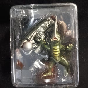May include: A green and yellow monster action figure with a long horn and sharp teeth, packaged in a clear plastic container. The figure has a detailed design and is posed in a dynamic stance.
