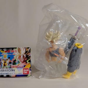 Puede incluir: Una figura de acción de Dragon Ball Z de Trunks en una bolsa de plástico, con una caja con varios personajes. La figura tiene cabello rubio puntiagudo, un atuendo azul y naranja y una espada púrpura. La caja tiene texto japonés y el logotipo de Bandai.