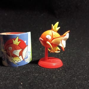 Könnte beinhalten: Eine orangefarbene und weiße Magikarp-Figur auf einem roten Ständer, neben einem blau-weißen zylindrischen Behälter mit einer Magikarp-Illustration. Die Figur hat eine gelbe Flosse und ist ein Sammlerstück.