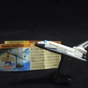 May include: A white and black toy space shuttle with "United States" on the side, displayed on a black stand. A box with Japanese text is in the background, showing a space shuttle illustration.