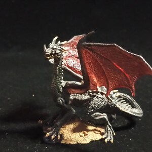 May include: A detailed figurine of a dragon with black scales and red wings. The dragon is posed on a small, tan base, with its head turned to the side. The wings are spread, and the dragon has a menacing expression.