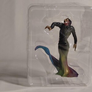 May include: A figurine of a person in a long, flowing gown with a rainbow gradient. The figure is in a dramatic pose, with arms outstretched and mouth open. The gown has a long, flowing train with blue, green, yellow, and purple hues.