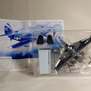 Op de afbeelding: Een modelbouwpakket van een SB2C Helldiver-vliegtuig in een transparante plastic verpakking. Het donkerblauwe en witte model is gedeeltelijk geassembleerd. De verpakking bevat een afbeelding van het vliegtuig en de tekst "SB2C Helldiver".