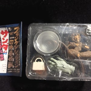 Könnte beinhalten: Ein Vintage-Actionfiguren-Set mit einer braunen, pelzigen Kreatur, einer grünen Hand und einem kleinen weißen Objekt, alles in einem transparenten Kunststoffbehälter. Eine blau-weiße japanische Textkarte ist ebenfalls enthalten.