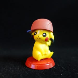 Peut inclure: Figurine jaune de Pikachu portant une casquette rouge avec une visière bleue. La figurine est posée sur un socle rouge. Elle a des oreilles noires, des joues rouges et un visage souriant.