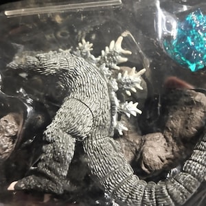 May include: A Godzilla action figure in a clear plastic package. The figure is primarily gray with white dorsal fins and a textured skin. A blue, crystal-like object is also visible.