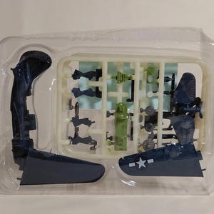 Puede incluir: Un kit de modelo de avión con varias piezas de plástico azul oscuro y verde. El kit incluye alas, una cola y otros componentes, todos empaquetados en un recipiente de plástico transparente. Las alas tienen un emblema de estrella blanca.