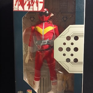 Puede incluir: Figura de acción vintage de un superhéroe rojo en una caja. La figura lleva un traje rojo con detalles amarillos, un casco y botas blancas. La caja tiene texto japonés y una ventana de plástico transparente.