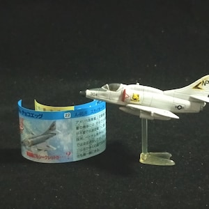 Pode incluir: Um modelo de avião branco e cinza, possivelmente um Skyhawk, é exibido em um suporte transparente. O modelo tem marcações pretas e amarelas e o número 304. Uma etiqueta curva azul e amarela com texto japonês está ao lado do avião.