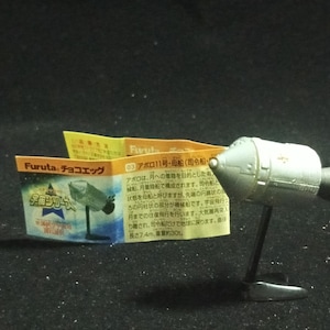 May include: A silver model of a spacecraft with a black base and a small, attached black cylinder. The model is accompanied by a small, folded paper with Japanese text and a graphic of the spacecraft.