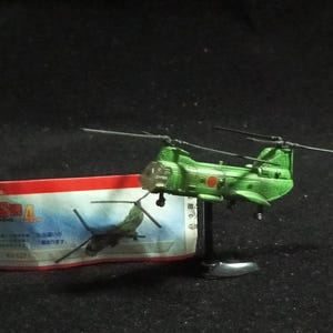 May include: A green toy helicopter with a red circle emblem and a silver stand. The helicopter has two rotors and is positioned next to a small box with Japanese text and a helicopter illustration.