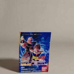 May include: A colorful Dragon Ball Z trading card pack featuring two characters in action poses against a blue background with white streaks. The pack has Japanese text and the Bandai logo.