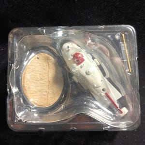 Peut inclure: Un modèle réduit d'hélicoptère blanc et rouge dans un emballage en plastique transparent. L'hélicoptère présente un design détaillé avec des accents rouges et une petite figurine rouge. L'emballage contient également une petite base en bois.