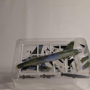 May include: A model airplane kit in a clear plastic container. The airplane is painted in camouflage green and blue. Additional parts, including missiles and wings, are also included in the kit.